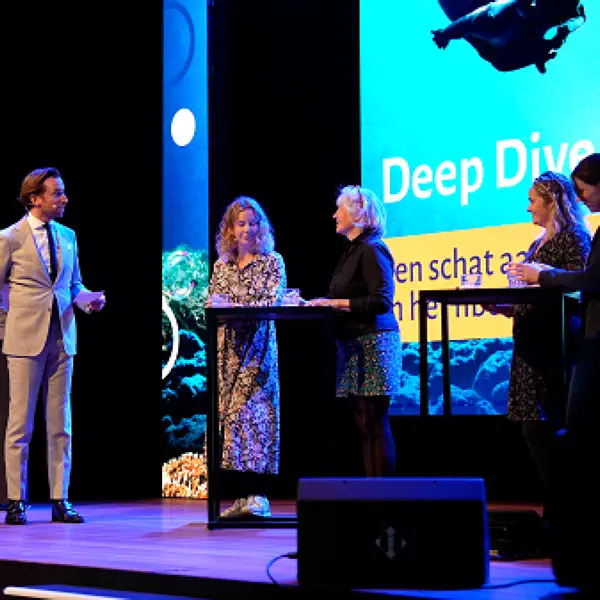 Foto van de paneldiscussie op een theaterpodium bij de Deep Dive van het NP Onderwijs: een man in een beige pak stelt een vraag aan vier mensen achter twee statafels. Achter hen een groot scherm met de omschrijving van de sessie bij de conferentie.