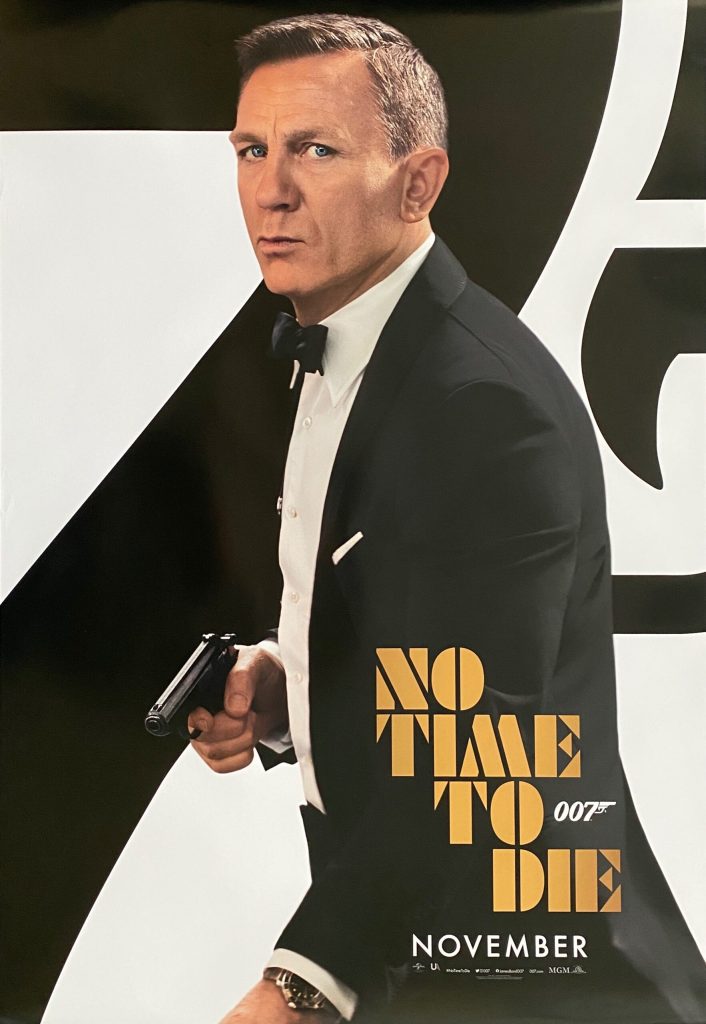 Filmposter van James Bond: No time to die. De 7 van 007 die is groter dan de hoogte van de poster is deels zichtbaar op de achtergrond in zwart. Daarvoor een foto van James bond en de titel in een retro lettertype.